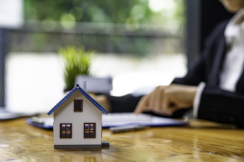 A model house sits on a desk. In the background, an attorney is completing legal paperwork for real estate law.